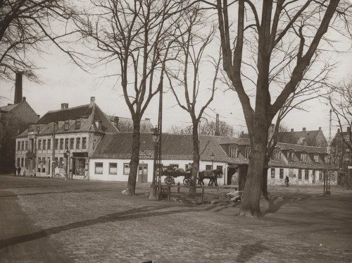 Gammelt fotografi af den lave kro Store Vibenshus bag store, nøgne træer, med en hestevogn foran og en fabriksskorsten i baggrunden.